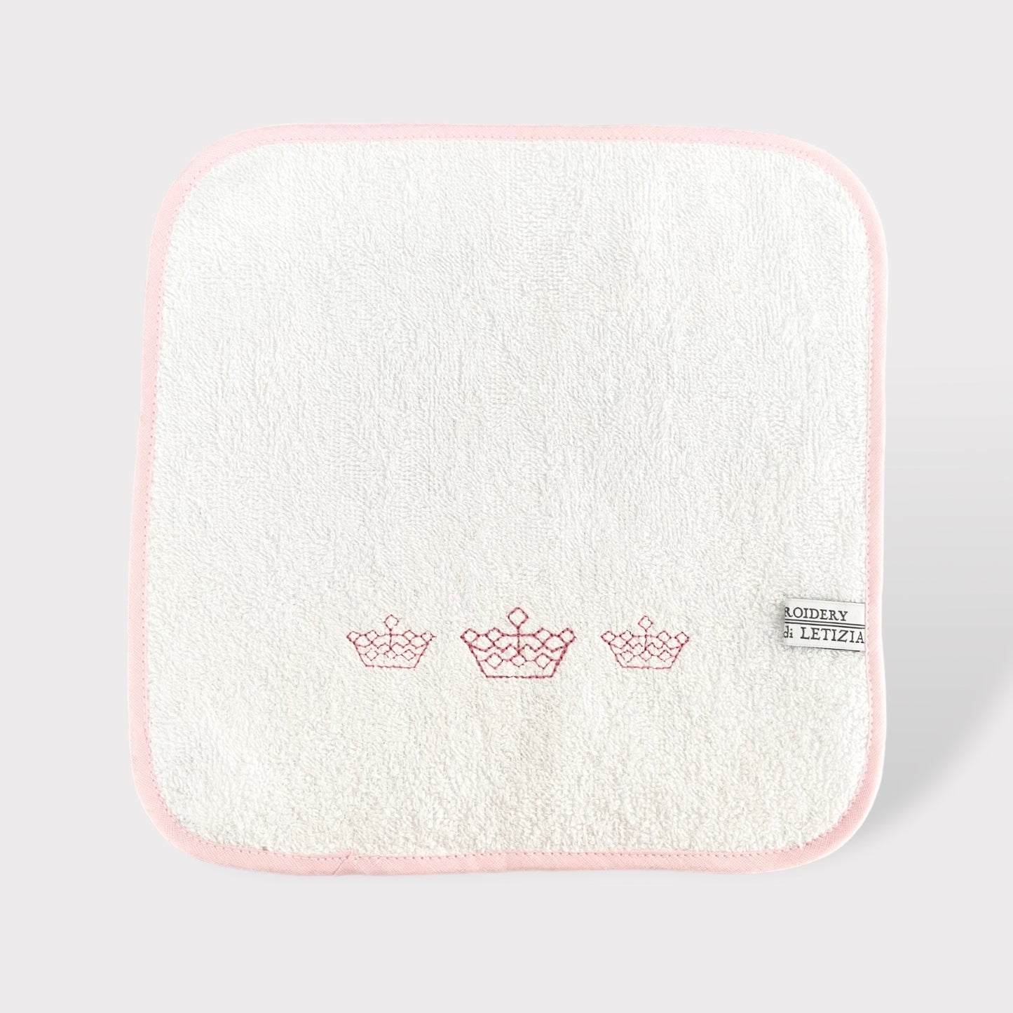 Asciuagamano in morbida spugna di cotone bianco con rifinitura rosa e ricamo di dolce corone da principessa.